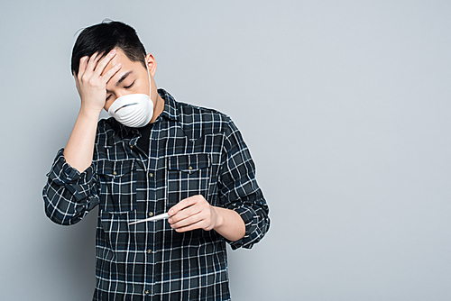young asian man in respirator mask looking at thermometer and touching forehead on grey background
