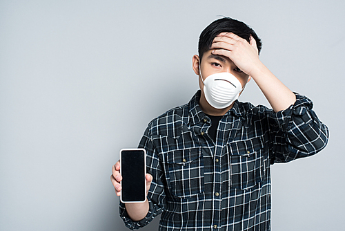young asian man in respirator mask touching forehead while showing smartphone with blank screen on grey background