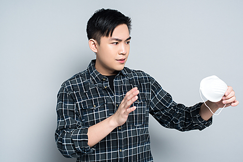 young asian man showing refuse gesture while holding respirator mask on grey background