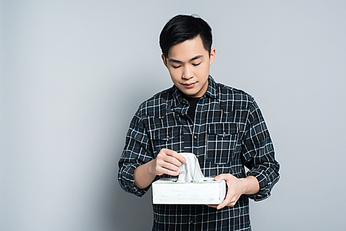young asian man holding pack of paper napkins while suffering from runny nose on grey background