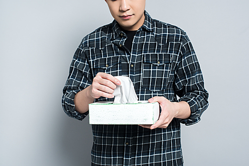 cropped view of young man holding pack of paper napkins while suffering from runny nose on grey background