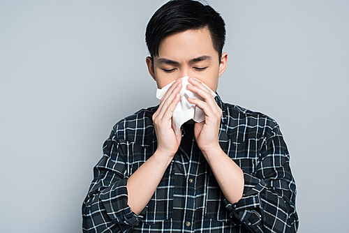 young asian man with closed eyes wiping nose with paper napkin while suffering from runny nose isolated on grey