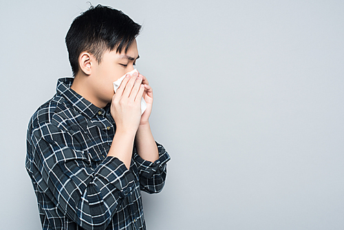 young asian man wiping nose with paper napkin while sneezing isolated on grey