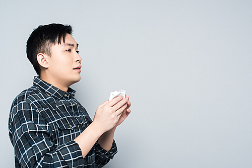 young asian man holding paper napkin while suffering from runny nose isolated on grey