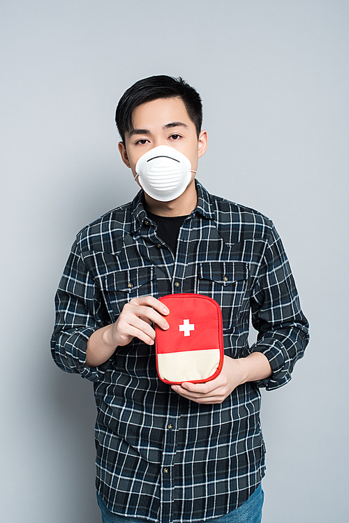 young asian man in respirator mask showing first aid kit while  on grey background