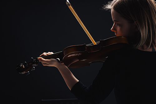 young concentrated woman playing cello with bow in darkness isolated on black