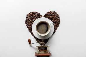 top view of coffee in cup on saucer with heart made of coffee grains near vintage coffee grinder on white background