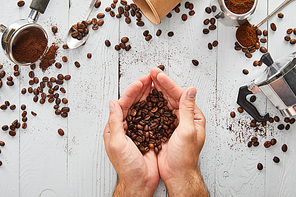 Top view of male hands with coffee beans under white wooden surface with spoons, paper cup, portafilter and geyser coffee maker