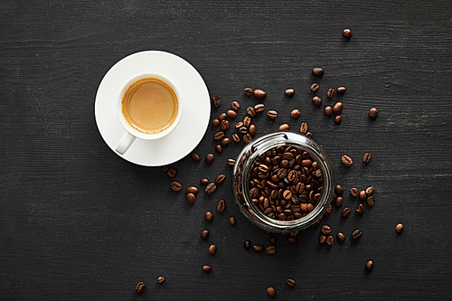 Top view of white cup of coffee on saucer near glass jar with coffee beans
