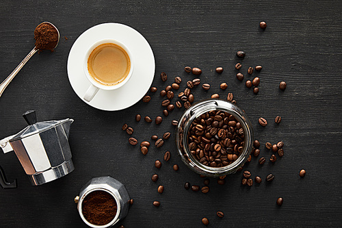 Top view of glass jar with coffee beans near geyser coffee maker, cup of coffee and spoon on dark wooden surface with coffee beans