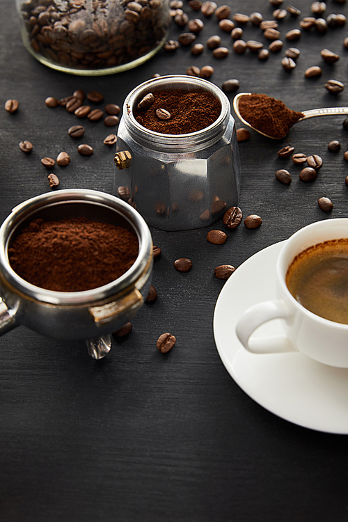 Part of geyser coffee maker near cup of coffee on saucer, portafilter and spoon on dark wooden surface with coffee beans