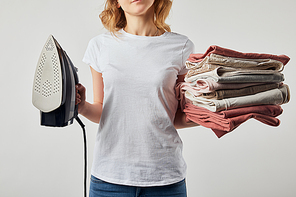 Partial woman in t-shirt holding iron and folded ironed clothes isolated on grey