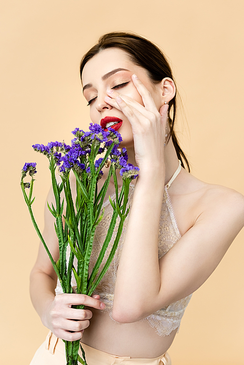 beautiful woman with closed eyes touching face and holding purple limonium flowers isolated on beige