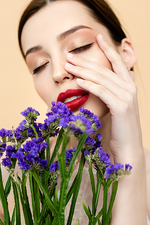 beautiful woman with closed eyes touching face near purple limonium flowers isolated on beige