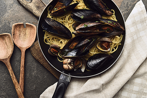 top view of delicious pasta with mollusks and mussels in frying pan on wooden cutting board near napkin and spatulas