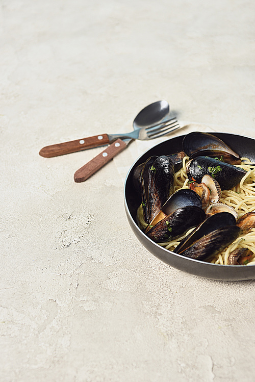 tasty pasta with seafood in frying pan near cutlery on textured grey background