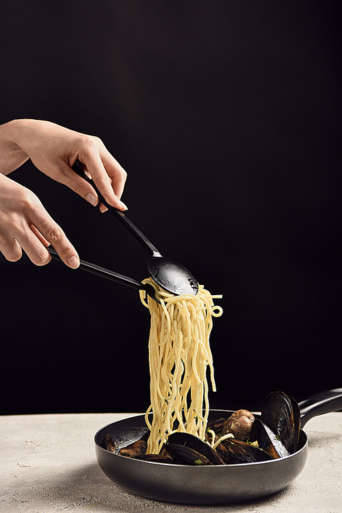 partial view of woman taking delicious Italian spaghetti with seafood from frying pan isolated on black