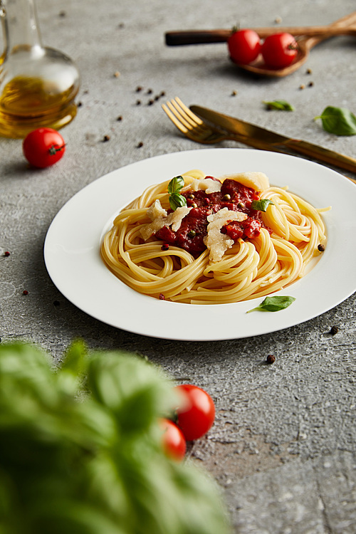 selective focus of tasty bolognese pasta with tomato sauce and Parmesan on white plate near ingredients and cutlery on grey background