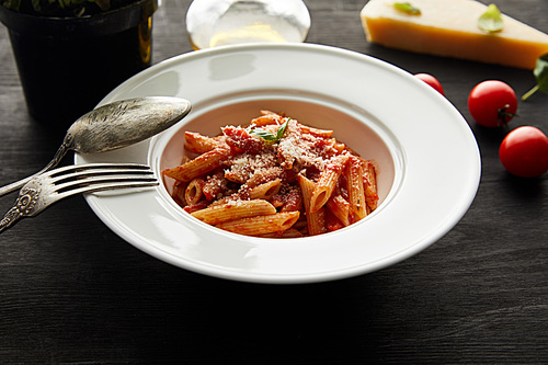 selective focus of tasty bolognese pasta with tomato sauce and Parmesan in white plate near ingredients and cutlery on black wooden background