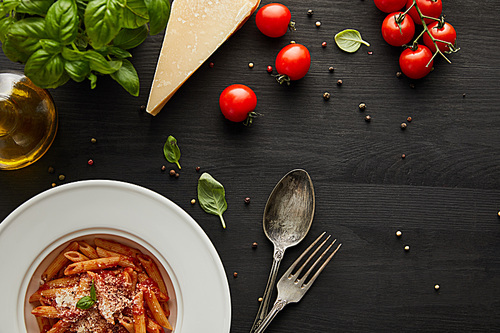 top view of tasty bolognese pasta with tomato sauce and Parmesan in white plate near ingredients and cutlery on black wooden background