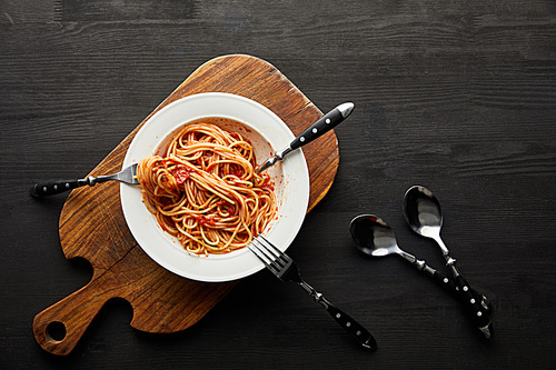 top view of tasty bolognese pasta with tomato sauce in white plate on wooden cutting board near cutlery on black wooden background