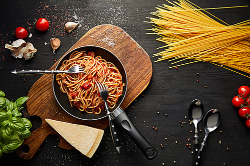 top view of tasty bolognese pasta in frying pan near ingredients and cutlery on black wooden background