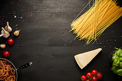top view of tasty bolognese pasta in frying pan near ingredients on black wooden background with copy space