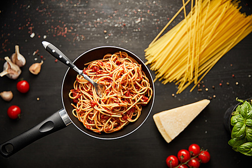 selective focus of tasty bolognese pasta in frying pan near ingredients on black wooden background