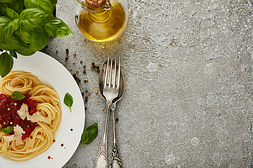 top view of delicious spaghetti with tomato sauce on plate near basil leaves, oil and cutlery on grey textured surface with copy space