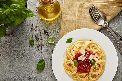 top view of delicious spaghetti with tomato sauce on plate near basil leaves, oil and cutlery on grey textured surface