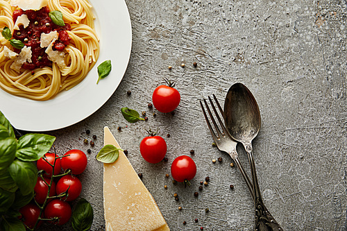 top view of delicious spaghetti with tomato sauce on plate near cheese, tomatoes and cutlery on grey textured surface