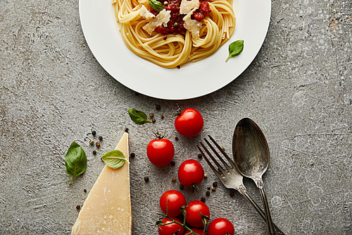 top view of delicious spaghetti with tomato sauce on plate near cheese, tomatoes and cutlery on grey textured surface