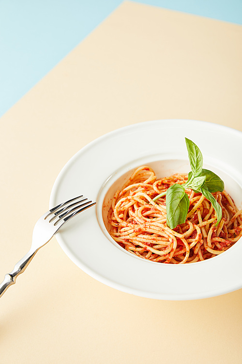 delicious spaghetti with tomato sauce in plate near fork on blue and yellow background