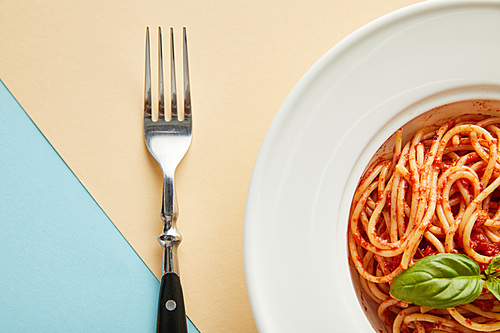 top view of delicious spaghetti with tomato sauce in plate near fork on blue and yellow background