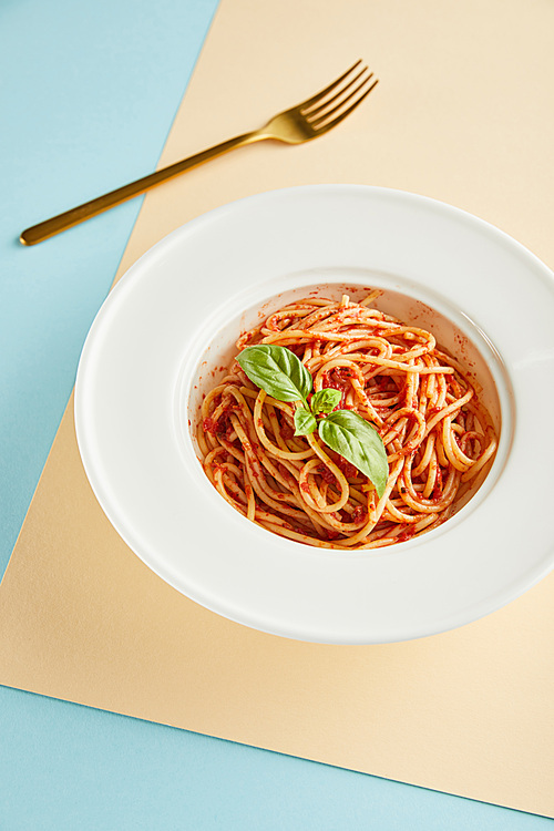 delicious spaghetti with tomato sauce and basil in plate near fork on blue and yellow background