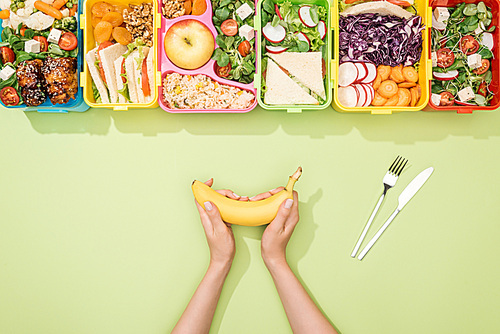 cropped view of woman holding banana near fork, knife and lunch boxes with food