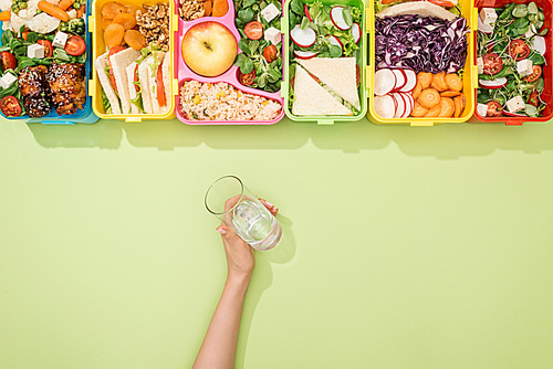 cropped view of woman holding glass of water in hand near lunch boxes with food