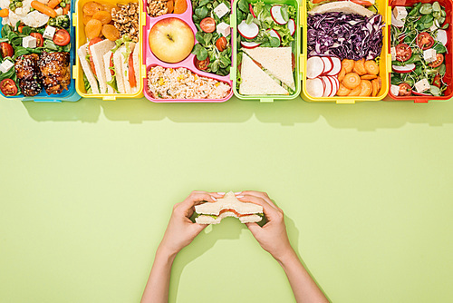 cropped view of woman holding sandwich in hands near lunch boxes with food