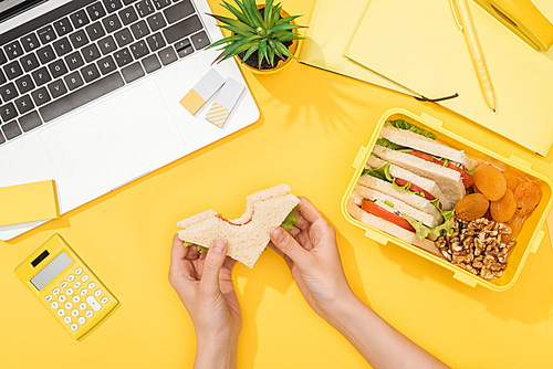 cropped view of woman holding sandwich in hands near lunch box, laptop and office supplies