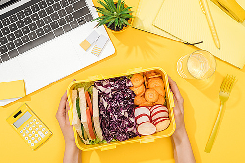 cropped view of woman holding lunch box near laptop and office supplies