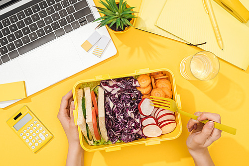 cropped view of woman holding lunch box with food near laptop and office supplies