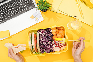 cropped view of woman holding sandwich in hand near lunch box with food, laptop, glass of water and office supplies