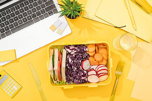 top view of lunch box with food near laptop, glass of water and office supplies
