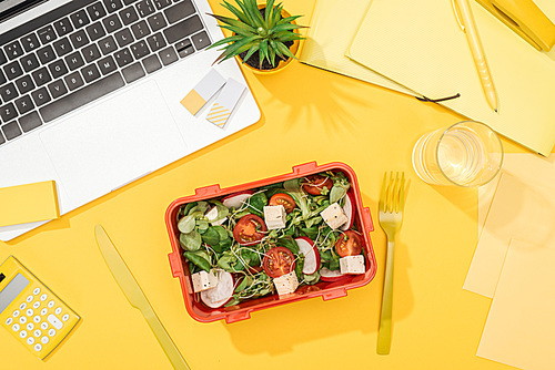 top view of lunch box with food near laptop, glass of water and office supplies
