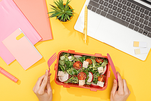 cropped view of woman holding fork over lunch box with food near laptop and office supplies
