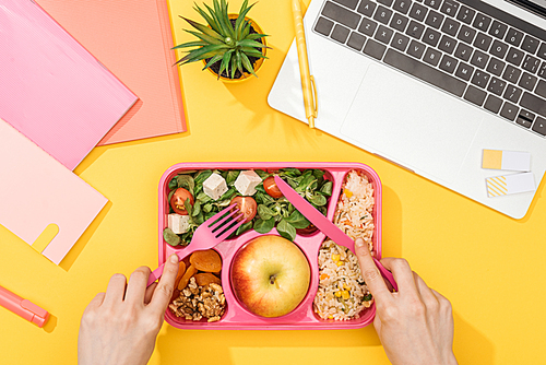 cropped view of woman holding fork over lunch box with food near laptop and office supplies
