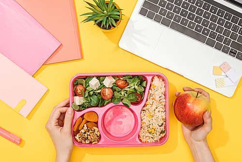 cropped view of woman holding lunch box with food near laptop and office supplies