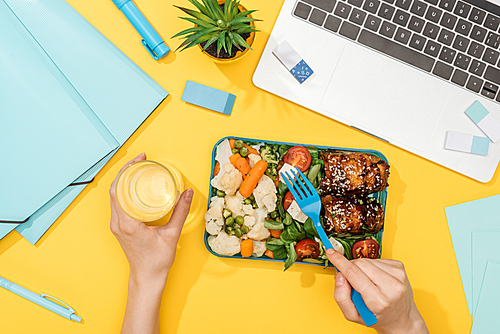 cropped view of woman holding lunch box with food and glass of water near laptop and office supplies