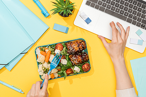 cropped view of woman working with laptop near lunch box and office supplies