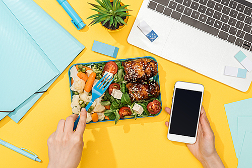 cropped view of woman holding smartphone near lunch box, laptop and office supplies